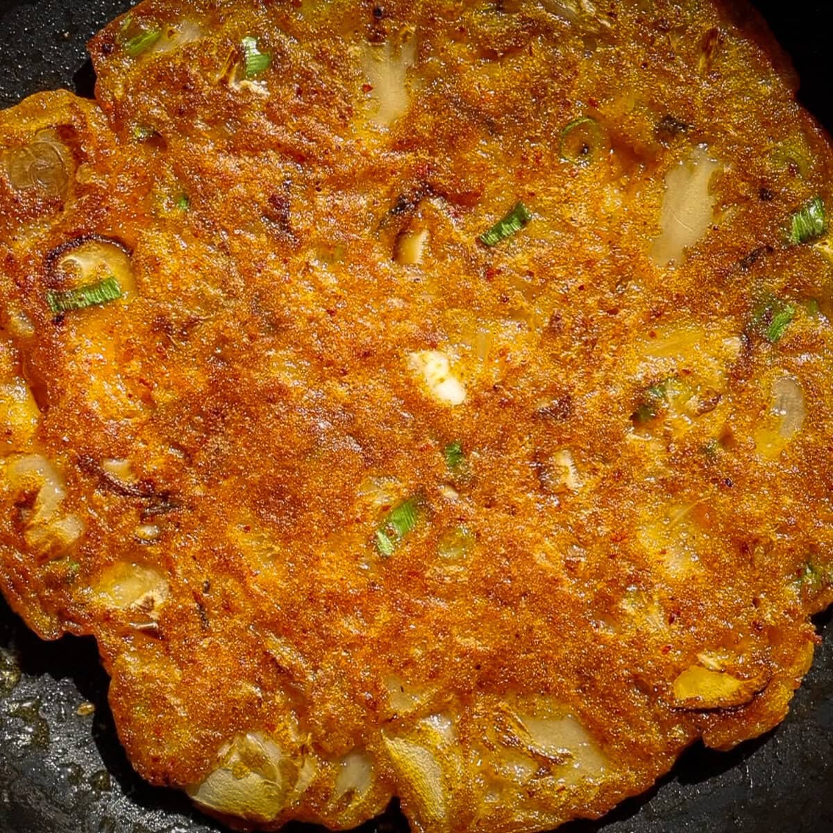 A close-up of golden-brown, crispy Korean kimchi pancakes with visible pieces of green onion and other vegetables cooked into the surface.